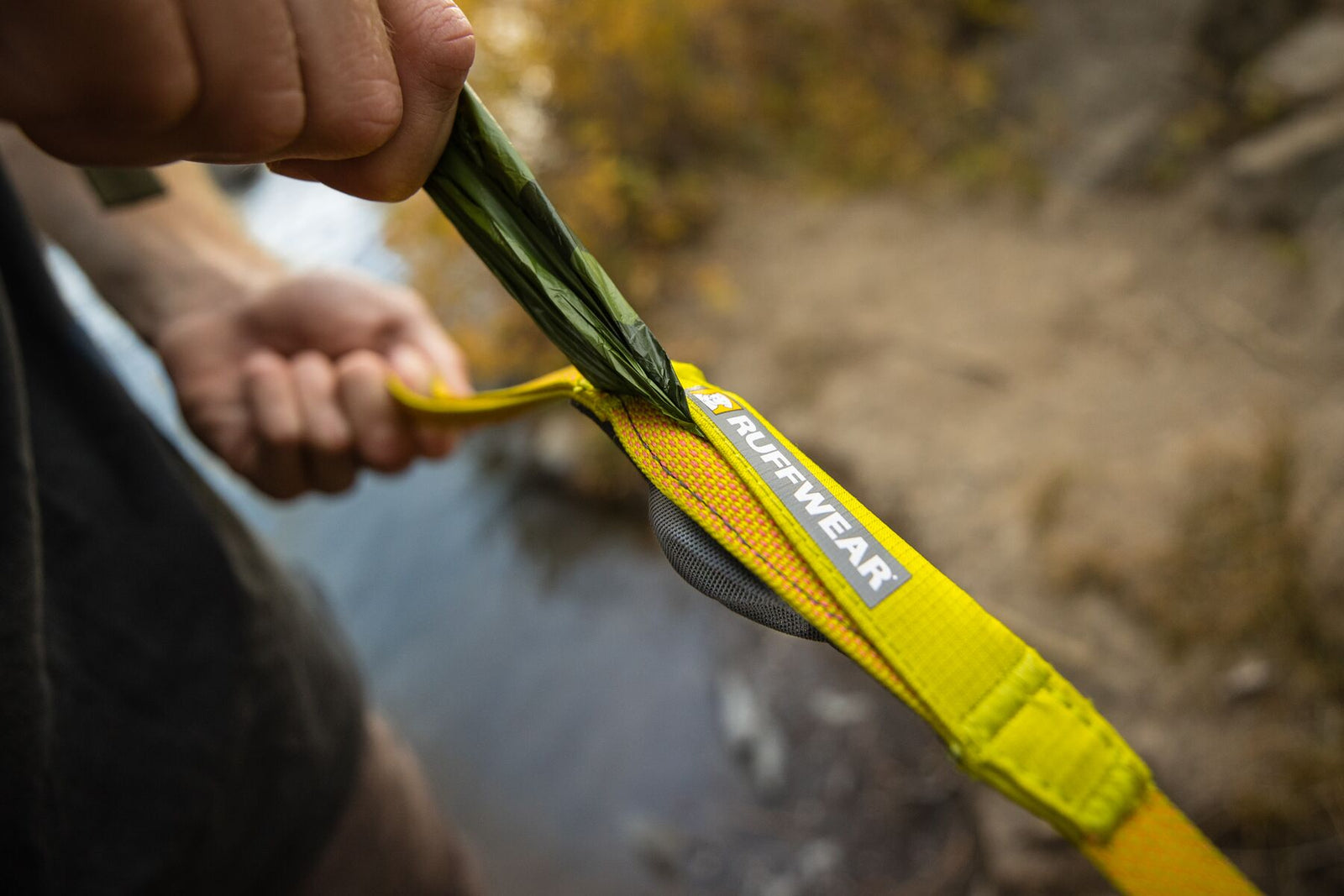 Close-up of a hand pulling a pick-up bag from the stash pocket on the Ruffwear Hi & Light Dog Lead in Lichen Green, outdoors by a stream